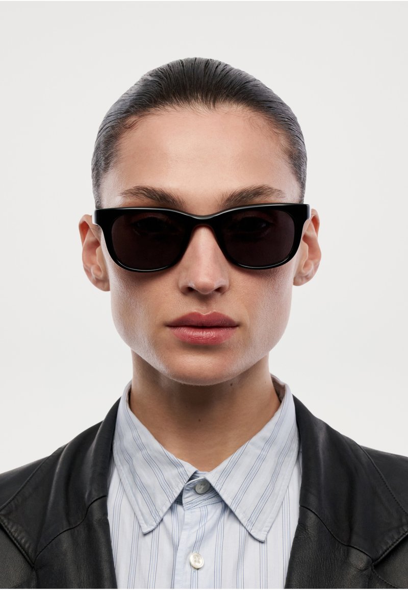 Personne aux cheveux gominés en arrière, portant des lunettes de soleil noires, une chemise rayée boutonnée et une veste en cuir noire devant un fond blanc uni.