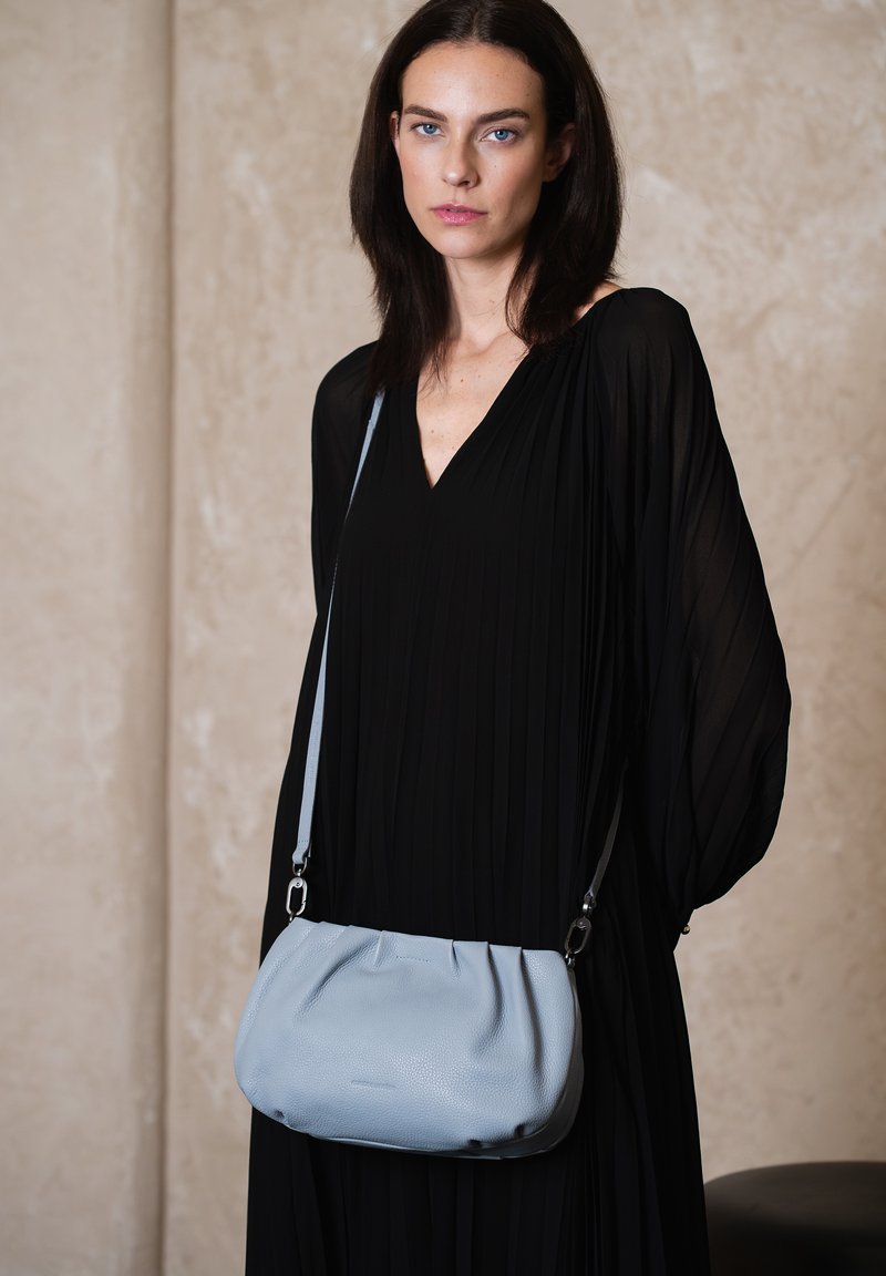 Hellblaues Leder-Crossbody-Bag mit einem gerafften Design, ausgestattet mit silbernen Beschlägen und einem langen verstellbaren Riemen, gehalten gegen ein schwarzes plissiertes Kleid.