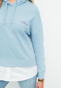 Sudadera azul claro con un bolsillo frontal, capucha con cordón y texto bordado "HISTORIAS DE NUEVA YORK" en púrpura. Tejido suave de mezcla de algodón.