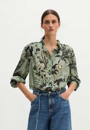 Frau trägt eine hellgrüne geblümte Bluse mit hochgekrempelten Ärmeln, in eine hoch taillierte blaue Jeans gesteckt, steht mit den Händen in den Taschen.