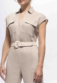 Žena u bež kombinezonu s kratkim rukavima i džepovima na prsima te širokim remenom u istoj boji, stoji nasuprot jednostavnoj svijetloj pozadini.