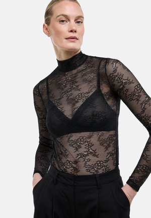 Femme blonde portant un haut à manches longues en dentelle noire à fleurs par-dessus un bralette noir, associé à un pantalon noir taille haute, les mains dans les poches.