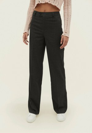 Frau trägt schwarze, hoch taillierte, weit geschnittene Hose mit Nadelstreifen, cropped beigen, abgenutzten Pullover und weiße Turnschuhe vor einfarbigem Hintergrund.