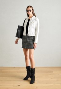 Veste en denim blanche, haut noir, mini-jupe à motifs et bottines en daim noir. Sac cabas noir et lunettes de soleil rondes visibles.