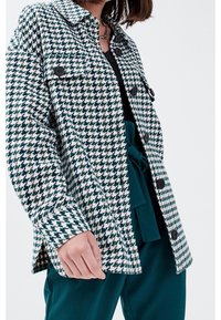 Groen-witte jas met pied-de-poule patroon, zwarte knopen, grote kraag en zijsplitten, gecombineerd met teal broek en een zwart topje.