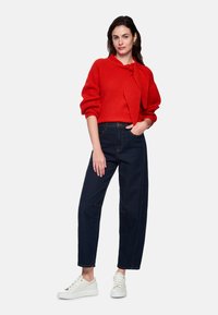 Roter Strickpullover mit einer Schleifendetail, kombiniert mit dunkelblauen Jeans mit weitem Bein und weißen Turnschuhen. Einfaches Design und strukturierter Stoff.