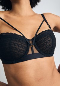 Black lace bra with sheer mesh panels, scalloped edges, and decorative straps. Features a central gold hardware accent.