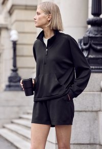 Tmavošedý fleecový pulóver na zips s vreckami v kombinácii s nohavicami krátkeho strihu v rovnakom odtieni. Hladký povrch. Čierny opakovane použiteľný pohár držaný v ľavej ruke.