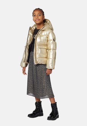 Doudoune dorée avec capuche, présentant une texture brillante et des poches zippées, associée à une jupe plissée à motifs et des bottes lacées noires.
