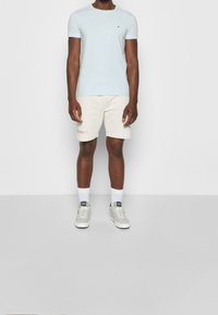 Hellblaues Baumwoll-T-Shirt mit kurzen Ärmeln, kombiniert mit cremefarbenen Shorts mit Logo-Print. Weiße Sportschuhe mit grauen Akzenten.