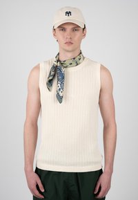 Jeune homme portant un pull sans manches côtelé crème, une écharpe verte à motif cachemire nouée au cou, une casquette beige avec un logo bleu marine, et un pantalon vert foncé.