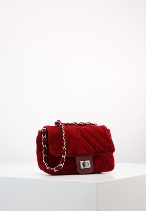 Rote, gestepptes Samt-Umhängetasche mit silberner Kettenriemen und rechteckigem silbernem Verschluss, auf weißem Podest vor schlichtem Hintergrund platziert.
