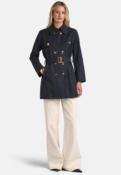 Trenci bleumarin cu nasturi aurii, talie cu curea și buzunare laterale; asortat cu pantaloni largi crem și pantofi cu vârf ascuțit.