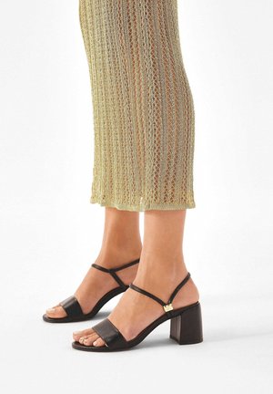 Füße, die schwarze Sandalen mit offenem Zehenbereich und Blockabsatz sowie dünnen Knöchelriemen tragen, kombiniert mit einem transparenten, goldfarbenen Strickkleid.