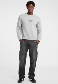 Šedá mikina s kulatým výstřihem a logem "Guess Jeans" na hrudi, v kombinaci s tmavě šedými cargo kalhotami s bočními kapsami a elastickým pasem.