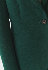 Tmavě zelený blejzr vyrobený z texturovaného materiálu, se zapínáním na jeden knoflík, klopami a boční kapsou.