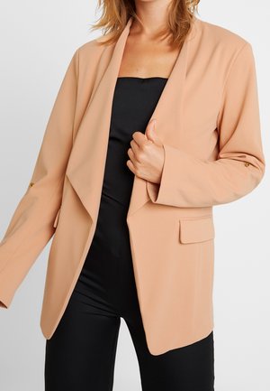Kvinde iført en beige blazer med åben front over et sort stropløst outfit, der holder blazerlappen med den ene hånd.