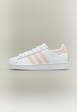 Λευκό αθλητικό παπούτσι Adidas Superstar με ροζ ανοιχτές ρίγες και ταμπελάκι στο τακούνι, με μυτερή μύτη και επίπεδη λαστιχένια σόλα, εμφανιζόμενο από το πλάι σε ουδέτερο φόντο.