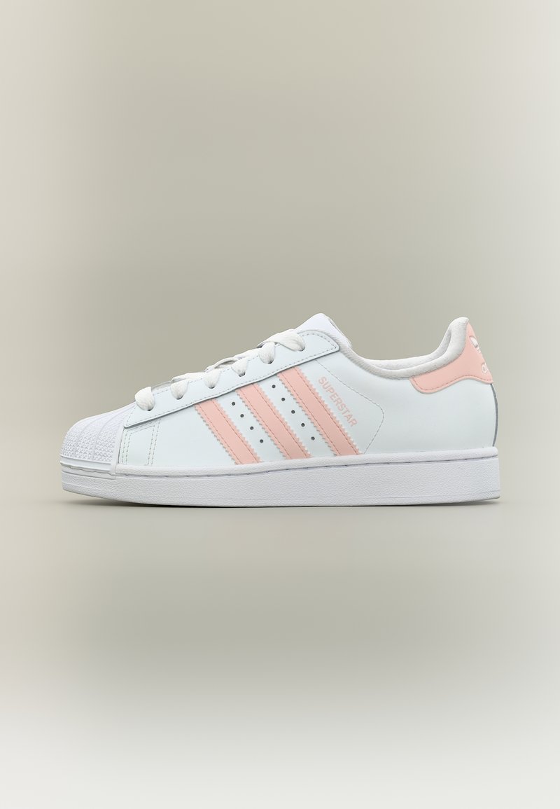 Λευκό αθλητικό παπούτσι Adidas Superstar με ροζ ανοιχτές ρίγες και ταμπελάκι στο τακούνι, με μυτερή μύτη και επίπεδη λαστιχένια σόλα, εμφανιζόμενο από το πλάι σε ουδέτερο φόντο.