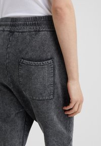 Närbild på en person som bär mörkgrå tvättade byxor med en bakfickapåse, höger arm avslappnat vid sidan mot en neutral bakgrund.