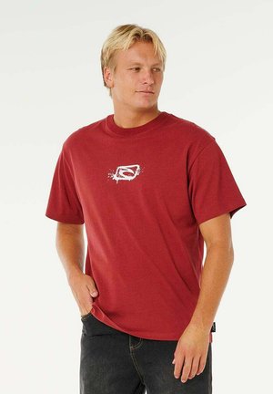 Blonde man draagt een rood T-shirt met een wit abstract logo, staat met één hand in zijn zak en kijkt opzij.