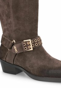 Botine maro din suede, până la genunchi, cu o suprafață texturată, având un cataramă decorativă din aur și o curea cu găici, și un toc mic, pătrat.