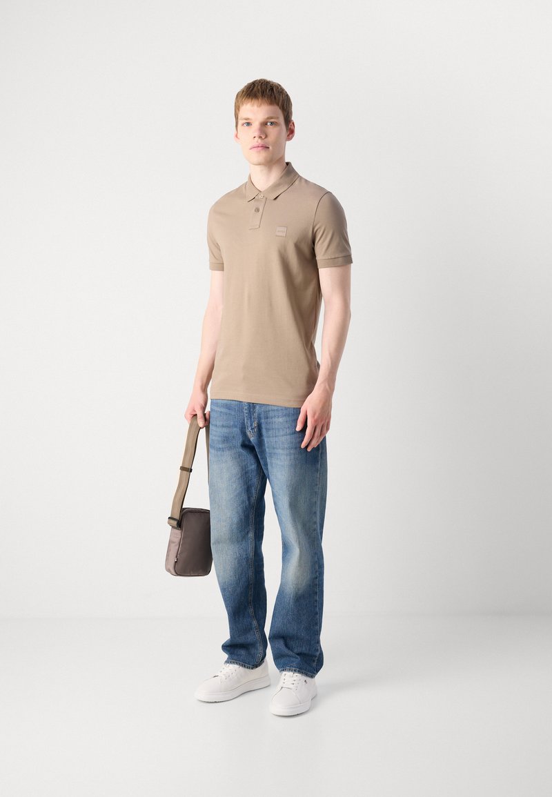 Polo-shirt à manches courtes marron clair, jeans bleus, baskets blanches et un petit sac à bandoulière marron. Le mannequin se tient contre un fond neutre.