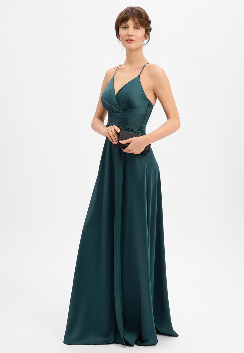 Femme en robe sans manches en satin bleu sarcelle tenant une pochette noire, debout devant un fond blanc uni.