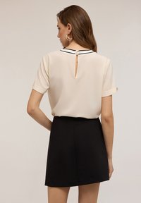 Blusa beige a maniche corte con dettaglio a chiave sul retro, abbinata a una mini gonna nera, con tessuto liscio e linee pulite.