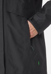 Giacca nera impermeabile con finitura testurizzata, mano in tasca con cordino regolabile e un piccolo dettaglio di logo verde.