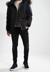 Personne portant un parka d'hiver noir avec capuche en fourrure, un pull en maille gris, un jean noir et des baskets bleues marine, debout avec une main dans la poche.