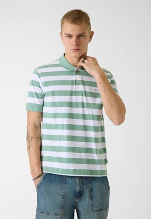 Młody mężczyzna z krótkimi blond włosami, ubrany w zielono-białą pasiastą koszulkę polo i niebieskie dżinsowe szorty, z tatuażami na ramionach, trzymający kołnierz.