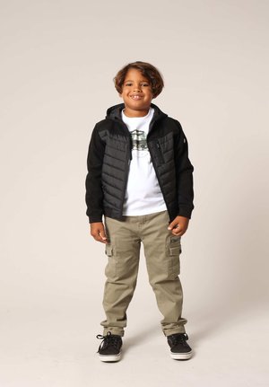 Veste noire zippée avec un devant matelassé, t-shirt blanc avec un motif graphique, pantalon cargo kaki, et baskets noires.