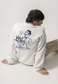 White sweatshirt featuring a blue graphic of a waiter with drinks and text "La Dolce Vita Kaotiko Society" on the back. Soft fabric, relaxed fit.