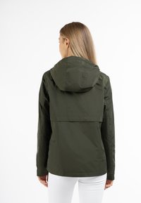 Giacca leggera verde oliva con cappuccio, dotata di una ventilazione posteriore e una texture liscia, abbinata a pantaloni bianchi.