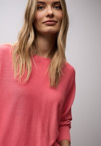 Jeune femme aux longs cheveux blonds portant un pull rose, debout devant un fond gris clair uni.