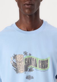 Lys blå bomull t-skjorte med et grafisk trykk av et tiki-ansikt, palmer, og teksten "SANTA CRUZ SKATEBOARDS" i svart og grønt.