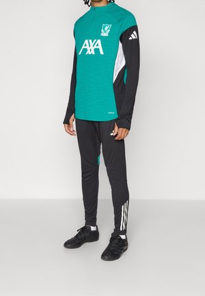 Męski model noszący długorękawową bluzę treningową Liverpool FC w kolorze morskim i czarnym oraz czarne spodnie dresowe Adidas z białymi paskami i czarne korki.