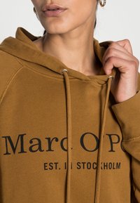 Felpa marrone in cotone, con stampa anteriore, cordini con accessori in argento e tasca a marsupio. Progettata con una vestibilità rilassata.