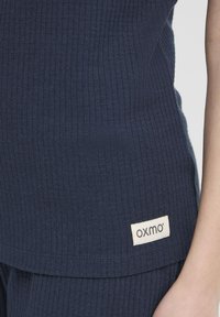 Canottiera a coste blu navy con un tessuto strutturato e una piccola etichetta color crema con il nome del brand "oxmo" cucita sul bordo inferiore.