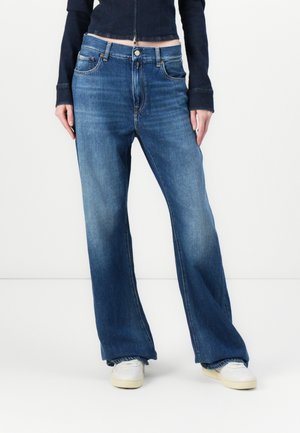 Persoon draagt blauwe wijde jeans, witte sneakers en een donker kort jasje, staand tegen een witte achtergrond.