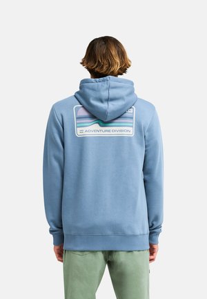 Persona che indossa una felpa con cappuccio azzurro chiaro con una grafica multicolore e il testo "Adventure Division" sulla schiena, abbinata a pantaloni verde chiaro.