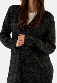 Kvinde med langt hår iført en sort og hvid tekstureret cardigan over en hvid skjorte, der justerer cardiganen med begge hænder.