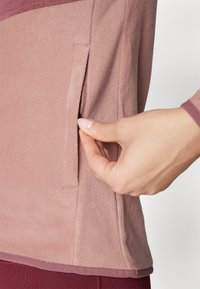 Maglione in morbido pile di un rosa tenue con tasca laterale; presenta una texture liscia e un dettaglio in orlo bordeaux a contrasto. Mano che si avvicina alla tasca.