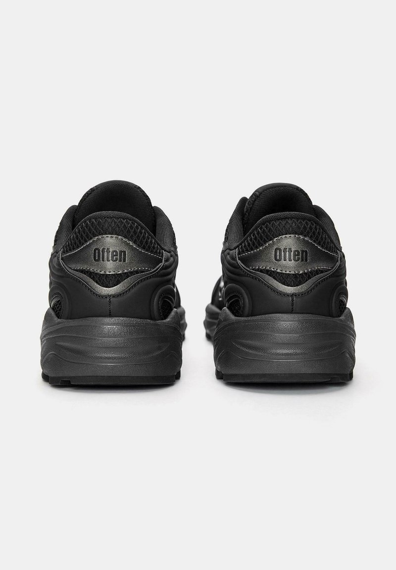 Une paire de chaussures de sport noires vue de dos avec la marque "Otten" sur l'étiquette au talon, sur un fond blanc.