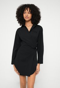 Čierne zavinovacie šaty s dlhými rukávmi, skladaným golierom a viazaním v páse. Na hrudi má jemný akcent s logom. Hladká textúra látky.