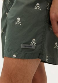 Șorturi de înot verzi cu un model alb de cranii și oase încrucișate. Prezintă un eticheta de marcă cu inscripția "SCALPERS" și are o textură netedă și ușoară.