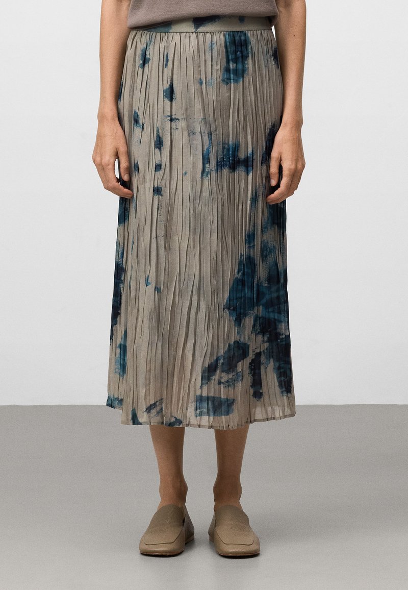 Vrouw in een geplooide midi rok met een blauw tie-dye patroon, taupe top en beige instappers, staande op een grijze vloer tegen een witte muur.