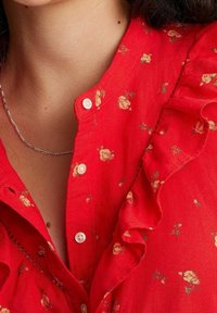 Chemise à boutons à fleurs rouges avec de petits motifs floraux dispersés, un col à volants et des détails de couture subtils.