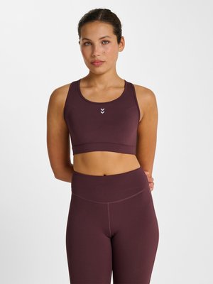 Kobieta z wiązanymi włosami, ubrana w ciemnofioletowy sportowy stanik i legginsy, stoi na białym tle, ręce za plecami.